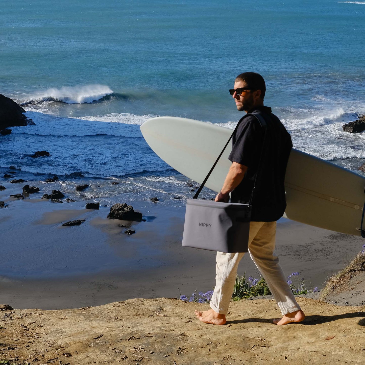 Stylish Grey NIPPY esky slung over shoulder of guy carying a surfboard at the beach showing how easy and light it is to carry