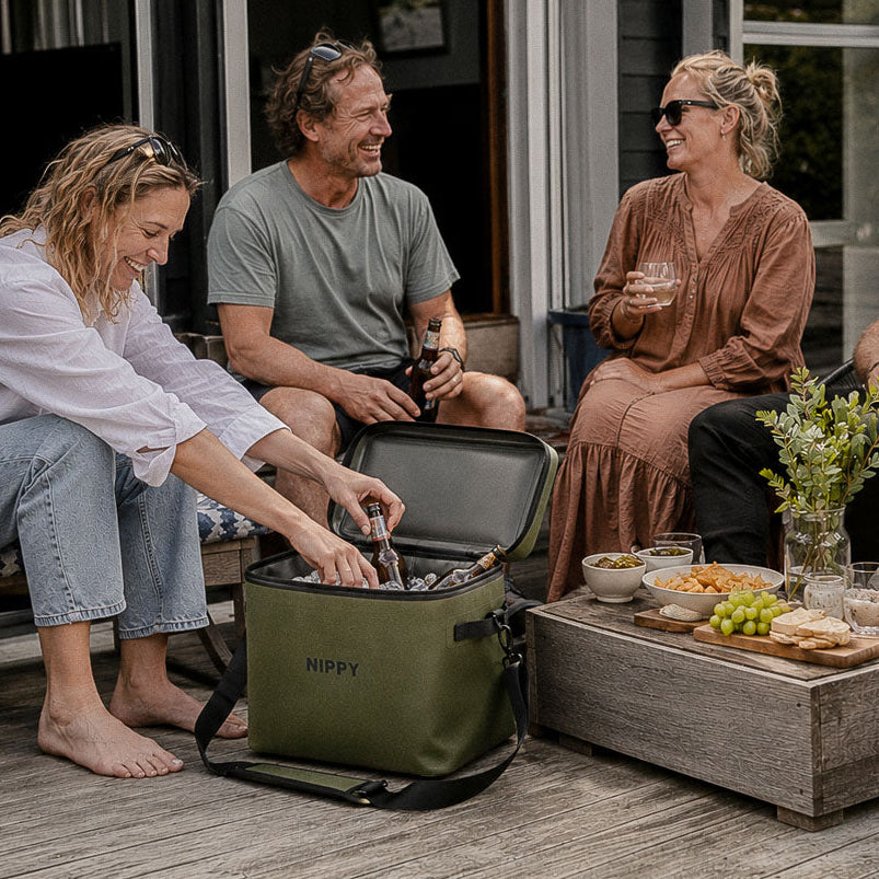 Four people sitting on a wooden deck around custom branded Nippy cooler they got as a staff gift. There is food and drinks on the coffee table in front of a modern house.