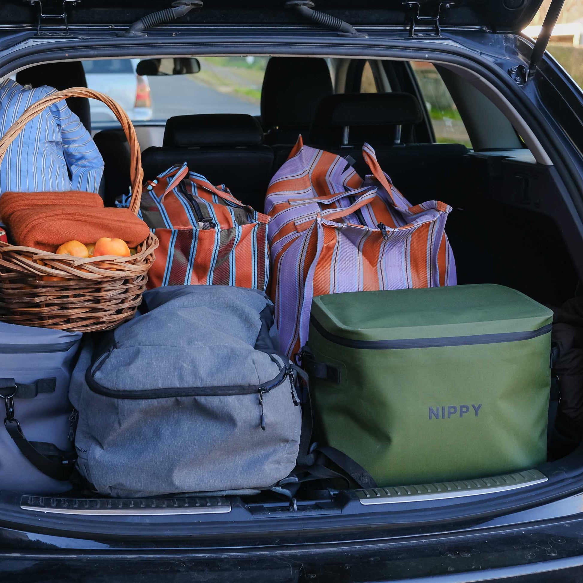 Trunk of a car filled with travel bags, an a NIPPY cooler, and a basket.