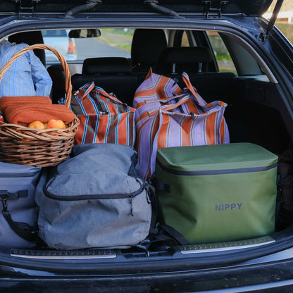 Trunk of a car filled with travel bags, an a NIPPY cooler, and a basket.